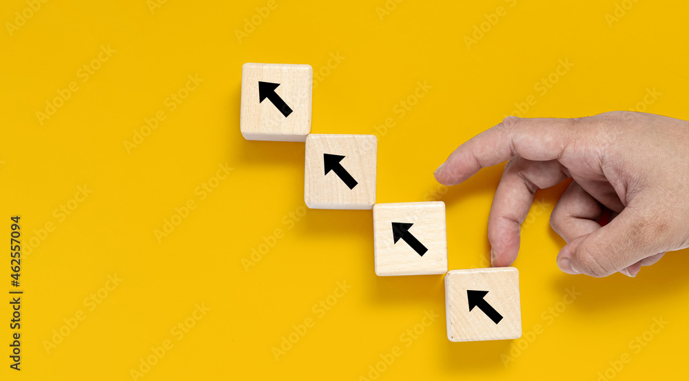 A square wooden block is placed on a yellow background, on the wooden block showing an up arrow and a hand doing an upward motion. banner with copy space for text, poster, mockup template. A square wooden block is placed on a yellow background, on the wooden block showing an up arrow and a hand doing an upward motion. banner with copy space for text, poster, mockup template.