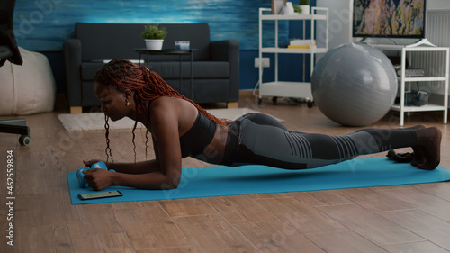 Flexible slim black woman stretching body muscles putting cronometer on phone practicing sport on yoga map during fitness workout in living room. Athetic adult enjoying healthy lifestyle photo