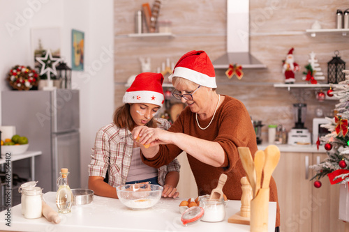 Grandmother showing to children how to prepare cookies dough breaking eggs in ingredients bowl enjoying winter holiday together in xmas decorated kitchen. Family celebrating christmas season