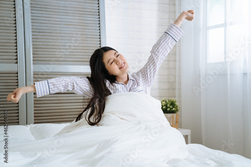 Woman stretching in bedroom after wake up. young asian korean beautiful lady sitting in comfort white bed smiling enjoy morning with sunlight. happy charming female raised arms and hands joyful face.