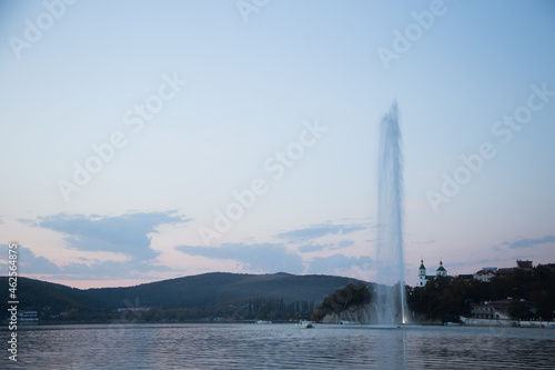 A fountain several meters high spouts from the center of the lake, you can see the mountains and the church, evening. Abrau Durso, Krasnodar Territory. High quality photo
