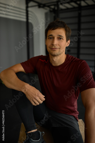 Attractive guy with cute face and muscular hands under sport shirt, sitting on wooden platform leaning hand on lifted bent knee, having calm, happy look, posing against gym wall with horizontal bars