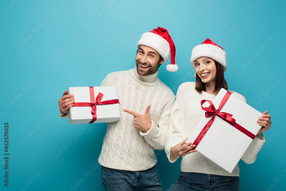 cheerful man in santa hat pointing at gift box near happy woman with present on blue cheerful man in santa hat pointing at gift box near happy woman with present on blue