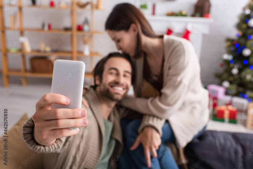 selective focus of smartphone in hand of happy man taking selfie with wife in decorated living room
