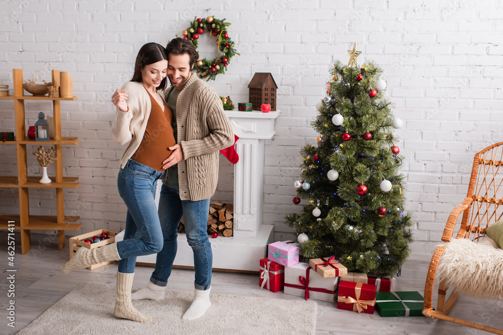 full length view of pregnant woman with happy husband near fireplace and christmas tree in living room full length view of pregnant woman with happy husband near fireplace and christmas tree in living room