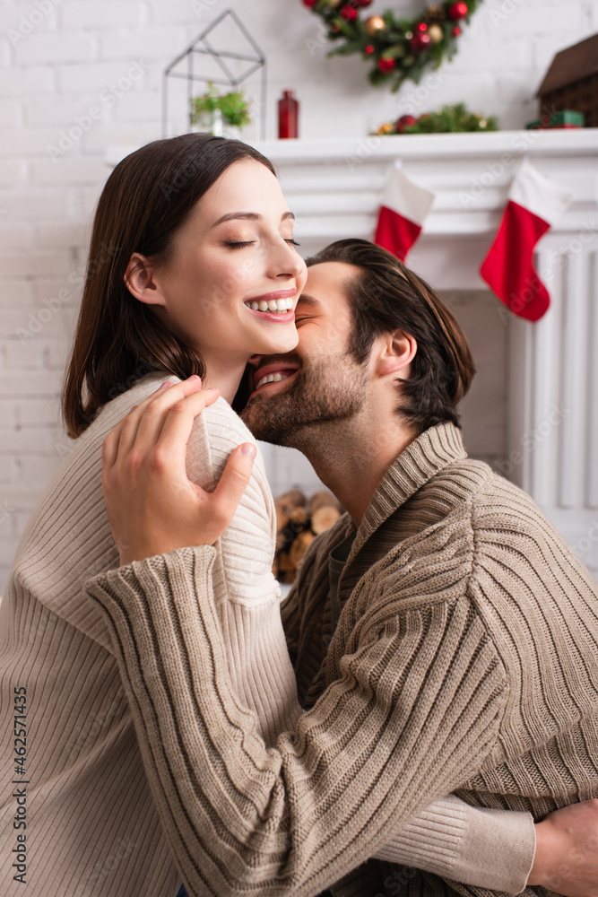 pleased man hugging wife near blurred fireplace with christmas decoration pleased man hugging wife near blurred fireplace with christmas decoration