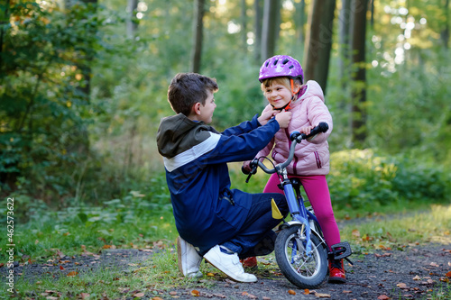 School kid boy, brother put on little preschool sister girl bike helmet on head. Brother teaching happy child cycling and having fun with learning bike. Active siblings family outdoors.