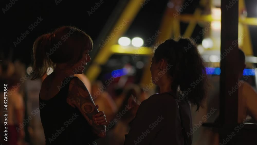Nightlife in amusement park. Two young women talking to each other. A glowing attraction with neon lights on the background. Carnival and holiday. Nightlife in amusement park. Two young women talking to each other. A glowing attraction with neon lights on the background. Carnival and holiday.