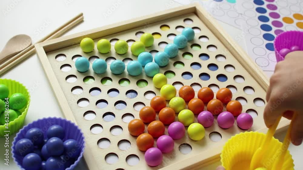 The child lays out colorful balls with tweezers. Wooden toy. Study of colors. Montessori concept, early brain exercise. Early education, fine motoric skills. Kids leisure. Preschool education. The child lays out colorful balls with tweezers. Wooden toy. Study of colors. Montessori concept, early brain exercise. Early education, fine motoric skills. Kids leisure. Preschool education.