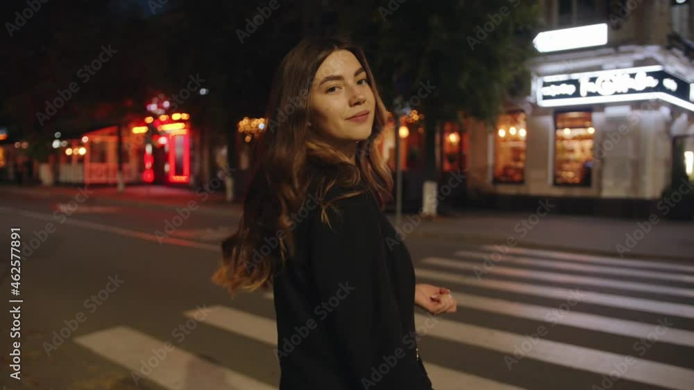Trendy young girl goes along a pedestrian crossing in the evening and heads to a summer cafe illuminated with neon lights and colorful garlands Trendy young girl goes along a pedestrian crossing in the evening and heads to a summer cafe illuminated with neon lights and colorful garlands