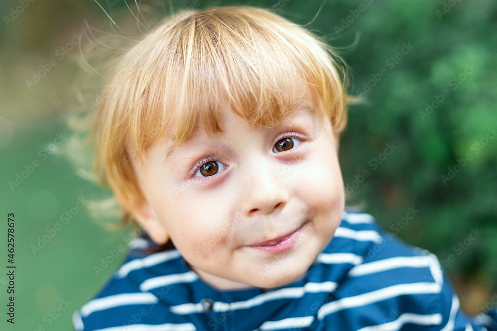 Close up portrait of a blond little boy outdoors. Child looking at the camera and smiling. Toddler wearing striped jacket spending time in a park in sunny fresh summer weather. Close up portrait of a blond little boy outdoors. Child looking at the camera and smiling. Toddler wearing striped jacket spending time in a park in sunny fresh summer weather.