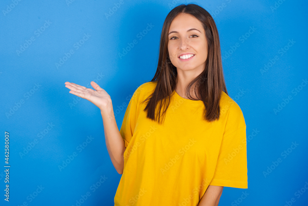 Positive glad Young european brunette woman wearing yellow T-shirt on blue says: wow how exciting it is, has amazed expression, shows something on blank space with open hand. Advertisement concept. Positive glad Young european brunette woman wearing yellow T-shirt on blue says: wow how exciting it is, has amazed expression, shows something on blank space with open hand. Advertisement concept.