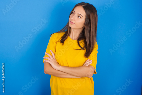 Image of upset Young european brunette woman wearing yellow T-shirt on blue background with arms crossed. Looking with disappointed expression aside after listening to bad news.