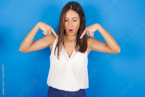 Amazed Young european brunette woman wearing white T-shirt on blue background points down with fore fingers, opens mouth being shocked. Advertisement concept. © Jihan
