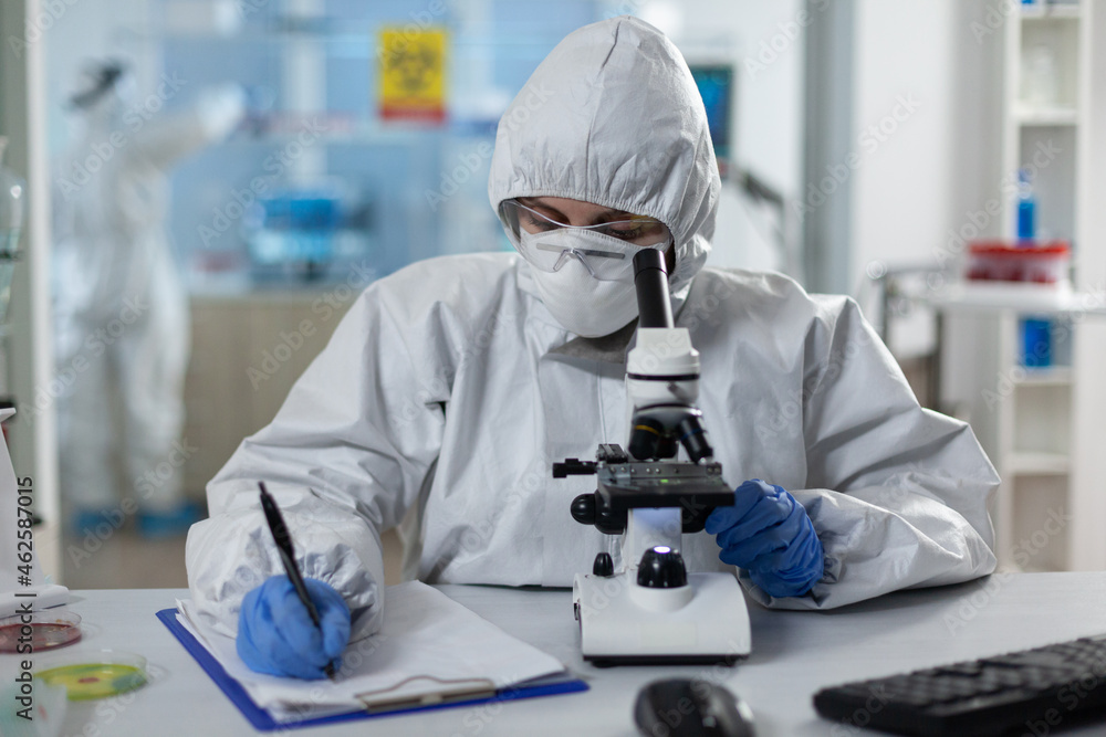 Biologist researcher wearing protective suit analyzing chemicals using medical microscope while writing clinical expertise during scientific experiment. Specialist doctor working in biochemistry lab Biologist researcher wearing protective suit analyzing chemicals using medical microscope while writing clinical expertise during scientific experiment. Specialist doctor working in biochemistry lab