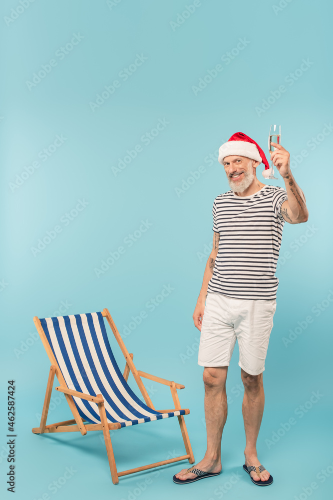 Obraz premium pleased mature man in santa hat holding glass of champagne near deck chair on blue