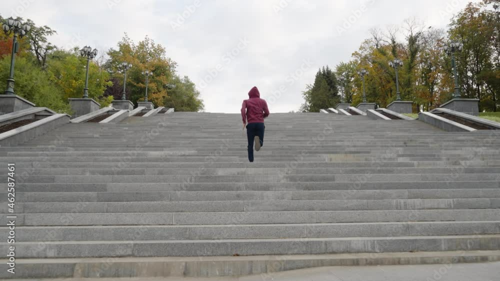 Slow motion fit man in sportswear with hood on head running up on stairs in park, yellow trees on sides. Following shot male athlete training outside on autumn morning. Concept of fitness Slow motion fit man in sportswear with hood on head running up on stairs in park, yellow trees on sides. Following shot male athlete training outside on autumn morning. Concept of fitness