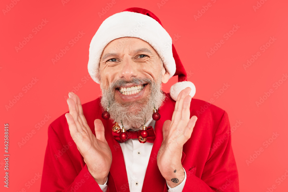 excited mature man in santa hat with christmas balls on beard gesturing isolated on red excited mature man in santa hat with christmas balls on beard gesturing isolated on red