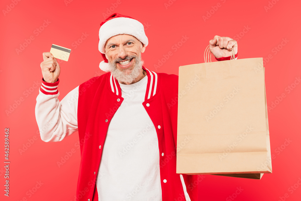 happy middle aged man in santa hat holding shopping bags and credit card isolated on red happy middle aged man in santa hat holding shopping bags and credit card isolated on red