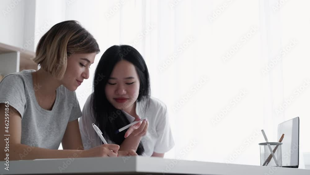College friends. Homework project. Classmate friendship. Distant learning. Joyful multiracial Asian Caucasian girls students buddies studying lesson at desk in light classroom.