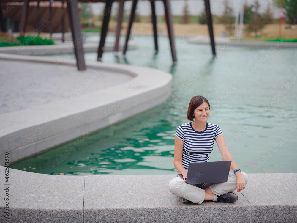 happy asian female student working with laptop in modern university campus park. woman in striped T-shirt and light trousers typing at gray laptop against the backdrop of a pond.