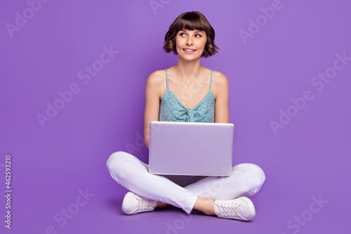 Portrait of attractive cheery minded girl sitting on floor using laptop thinking isolated over bright purple violet color background