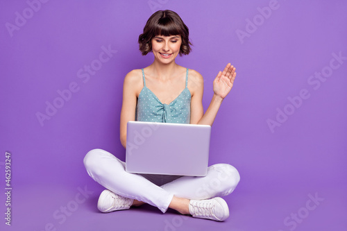 Portrait of attractive cheerful girl sitting using laptop calling waving hi hello isolated over bright violet purple color background