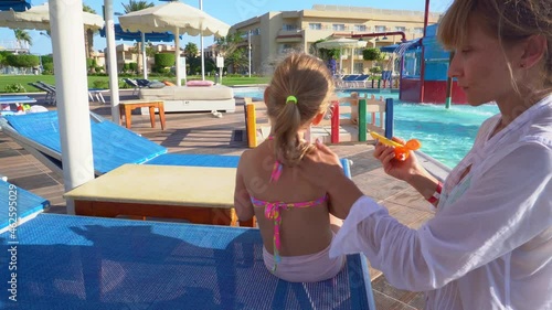 The child is sitting on a sun lounger near the pool. Mom puts sunscreen on the baby's back. 