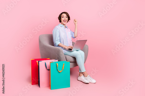 Full length body size photo woman sitting with packages working on computer gesturing like winner isolated pastel pink color background