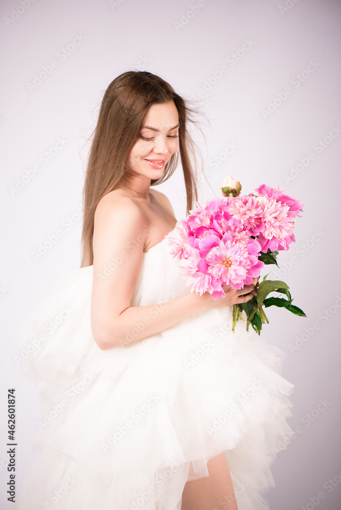 Beautiful cheerful young blonde girl wearing dress standing isolated over white background, holding bouquet of peonies