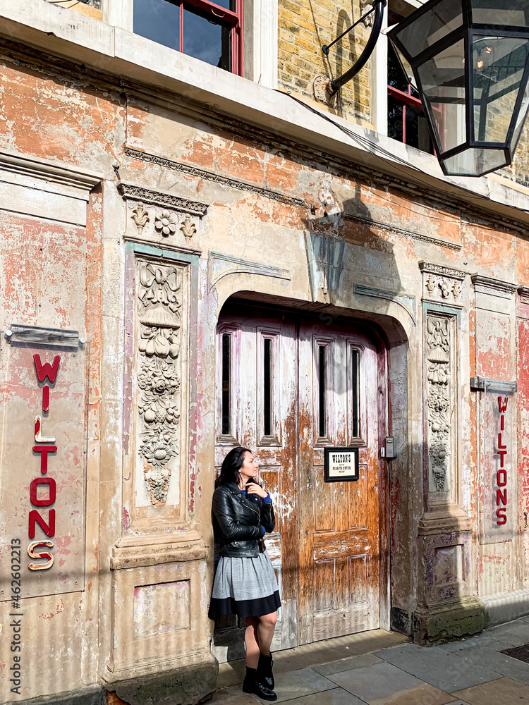 Young candid millennial woman stands by shabby wall. Young brunette girl in a dress, leather jacket and sunglasses stands by old shabby chic wall and door. Old house building facade. Old music hall