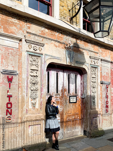 Young candid millennial woman stands by shabby wall. Young brunette girl in a dress, leather jacket and sunglasses stands by old shabby chic wall and door. Old house building facade. Old music hall © Alla
