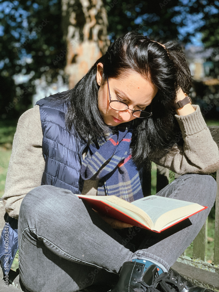 Young woman is reading red book in a park. Young candid millennial woman in casual autumnal clothes siting on a wooden bench in park and reading red book. Concept of back to school or university Young woman is reading red book in a park. Young candid millennial woman in casual autumnal clothes siting on a wooden bench in park and reading red book. Concept of back to school or university