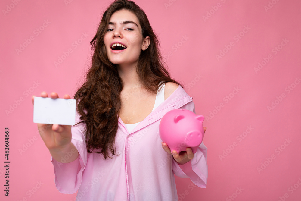 Beautiful happy joyful young brunette woman wearing shirt isolated on pink background with free space and holding pink pig money box and credit card for mockup Beautiful happy joyful young brunette woman wearing shirt isolated on pink background with free space and holding pink pig money box and credit card for mockup