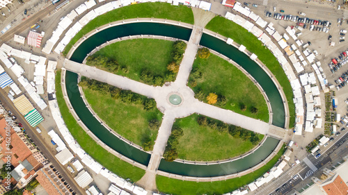 A huge oval square with green lawns, trees, a water channel and a cruciform path. View from above