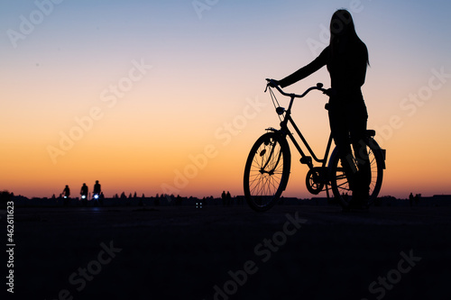 A silhouette of a young woman standing with female bicycle against sunset sky.