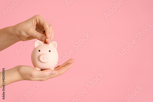 A woman s hand puts a coin in a pink piggy bank  copyspace on a pink background