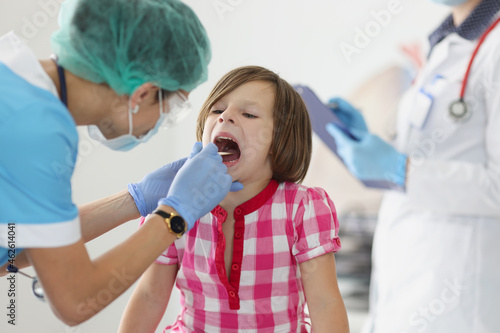 Otorhinolaryngologist doctor in protective medical mask examining throat of little girl using spatula in clinic photo