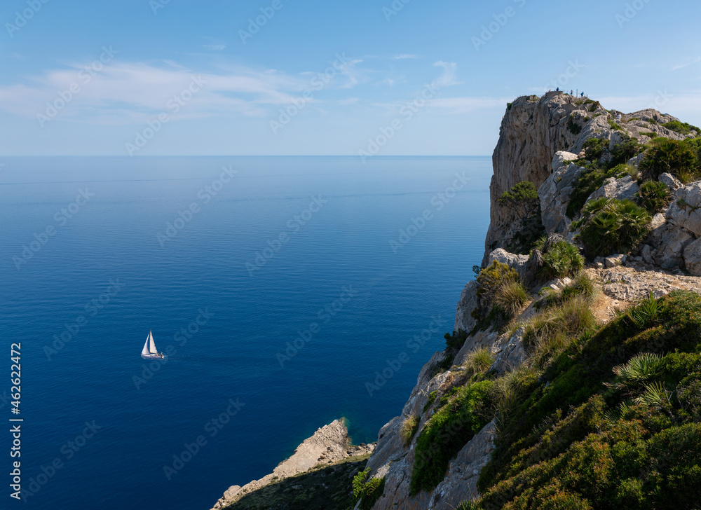 The steep cliffs of the Formentor peninsula in the east of the Mediterranean island of Mallorca. A sailing boat is sailing on the sea. The water is deep blue. Above is a viewing platform.