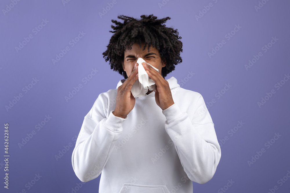 Cold and flu. Sick black teenager sneezing in paper tissue, suffering from rhinitis or sinusitis on violet background Cold and flu. Sick black teenager sneezing in paper tissue, suffering from rhinitis or sinusitis on violet background
