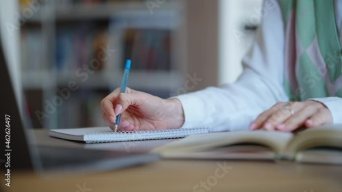 Female hand writing on paper, anoter hand flipping pages of book, bookshelves on blurred background. Closeup student sitting in library and studying. Concept of education photo