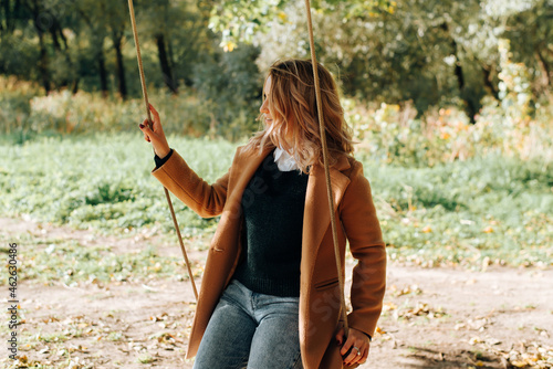 Profile portrait of young hipster woman in coat swinging, having fun on hanging tree swing, enjoying free time and leisure in nature. Beautiful smiling girl on seesaw outdoors, lifestyle #462630486