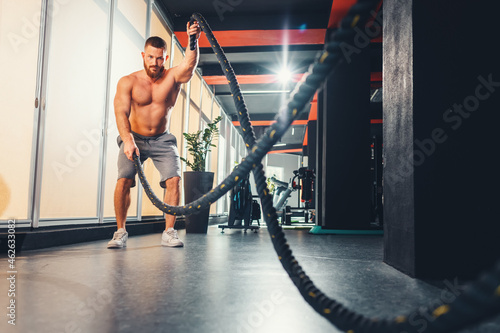 Athletic young man with battle rope doing exercise in fitness gym. Keeping fit with cross training