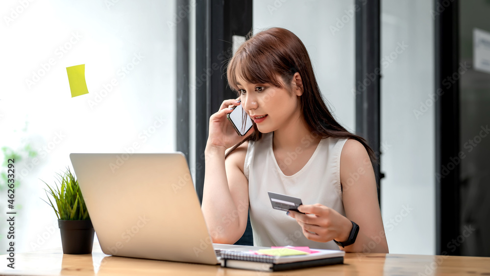 Beautiful young Asian businesswoman talking on the phone holding a credit card laptop at the office. Beautiful young Asian businesswoman talking on the phone holding a credit card laptop at the office.