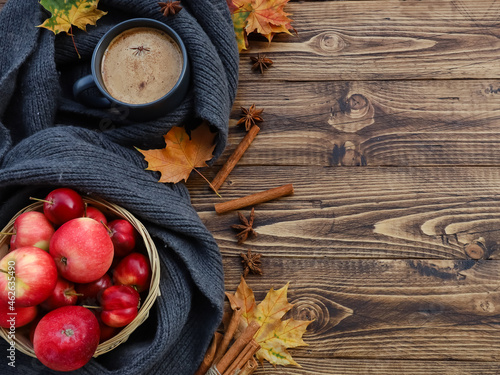 Latte mug wrapped in dark grey scarf, ripe red apples basket, fall, maple leaves, spices, cinnamon sticks top view on dark wooden table. Autumn hot drink, food concept