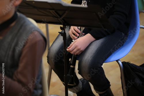 Student musician with a clarinet in lowered hands sitting on a chair at a rehearsal of a school orchestra