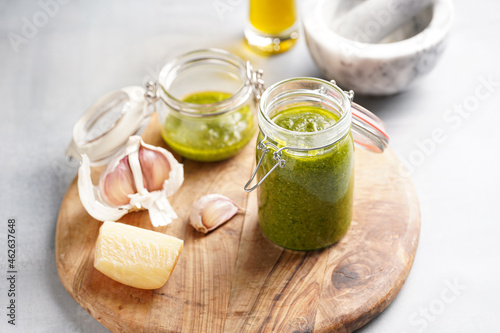 Several small glass mason jars with green italian basil sauce pesto on a round wooden tray with hard cheese pecorino and garlic cloves