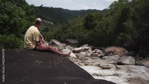 Young man tourist enjoys looking at forestry hills sitting on black rock near river rapids with huge sharp stones backside view photo