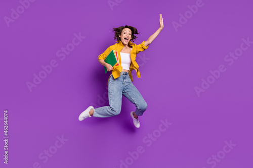 Photo of pretty excited young woman dressed yellow shirt smiling jumping high holding copybooks isolated purple color background