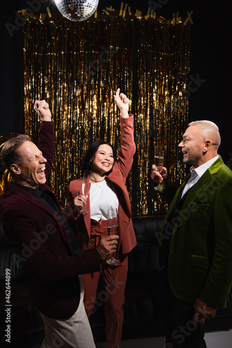 Excited multiethnic friends with champagne showing yes gesture near disco ball and tinsel on black background © LIGHTFIELD STUDIOS